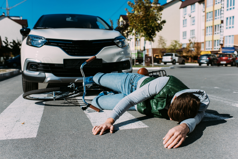 Residential city street with large apartment buildings: a young adult male is laying on the street in the crosswalk next to a bicycle, the man is face down and in a prone position. A large white SUV is stopped just in front of the man and the bicycle is partially underneath it.