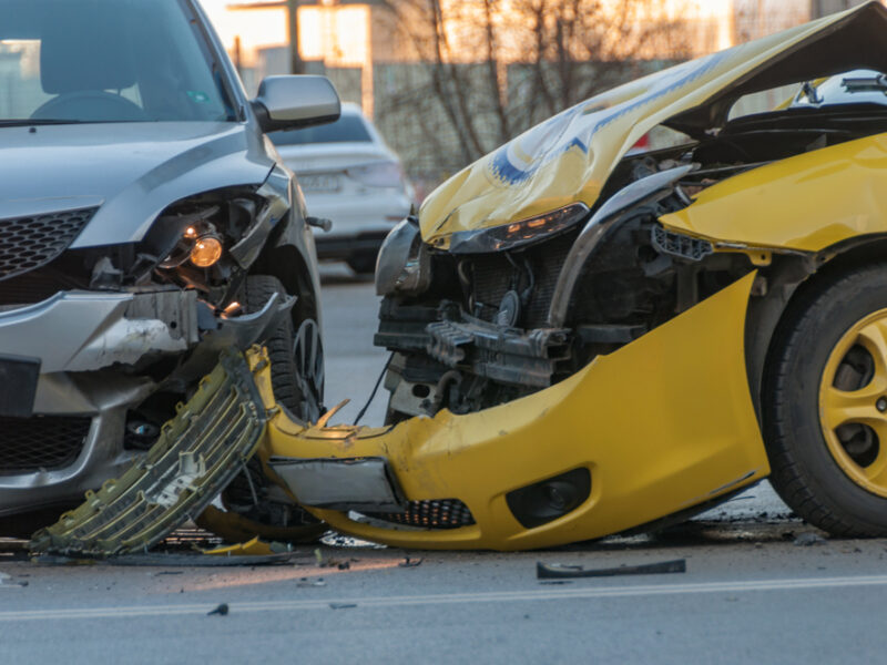 A yellow taxi cab has a smashed front end after a collision with a gray car on a city street