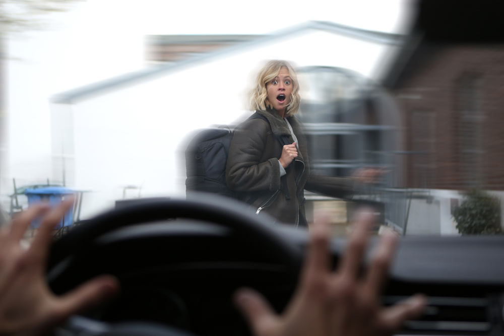 Shocked young blond woman who is crossing the street sees a car driving towards her, we can see the steering wheel and the driver's hands