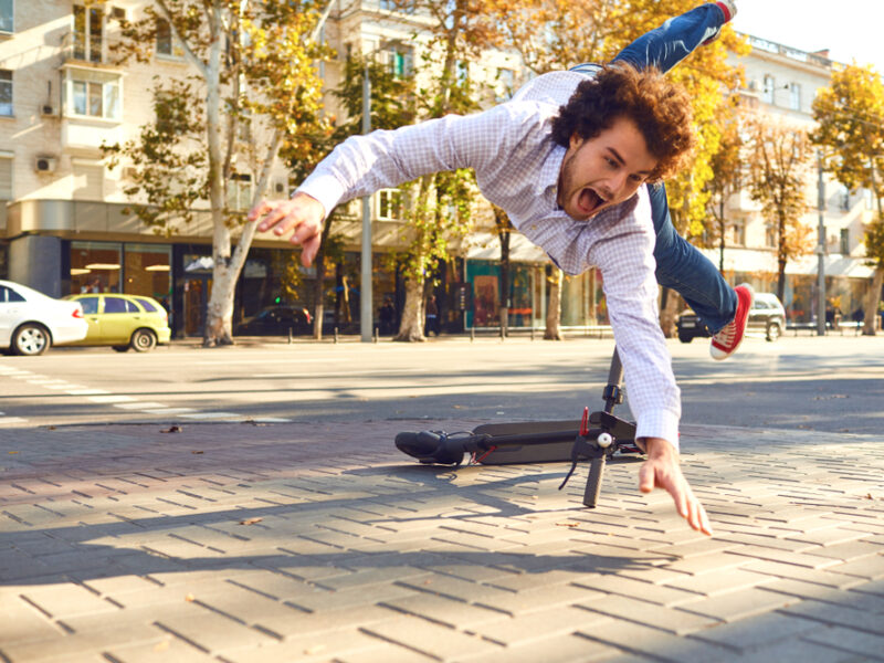 Man is mid-air and falling toward the sidewalk with his hands out to brace his fall. Behind him is an electric scooter that has turned on its side.