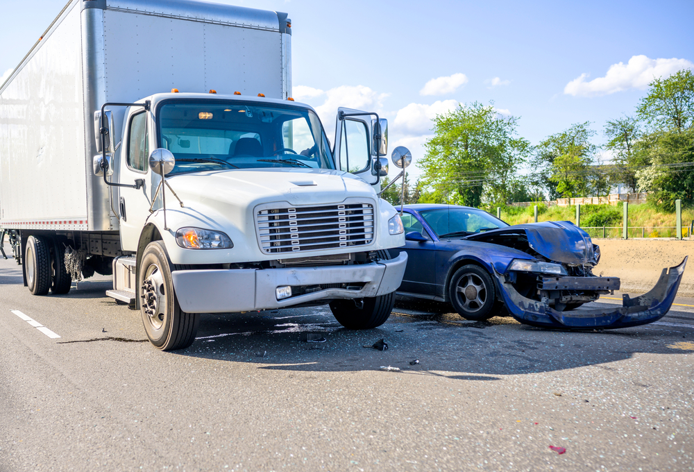 A semi box truck shows front bumper damage as it is stopped on the road next to a demolished blue car with the entire front bumper ripped off and laying in front of the vehicle's smashed hood and front end.