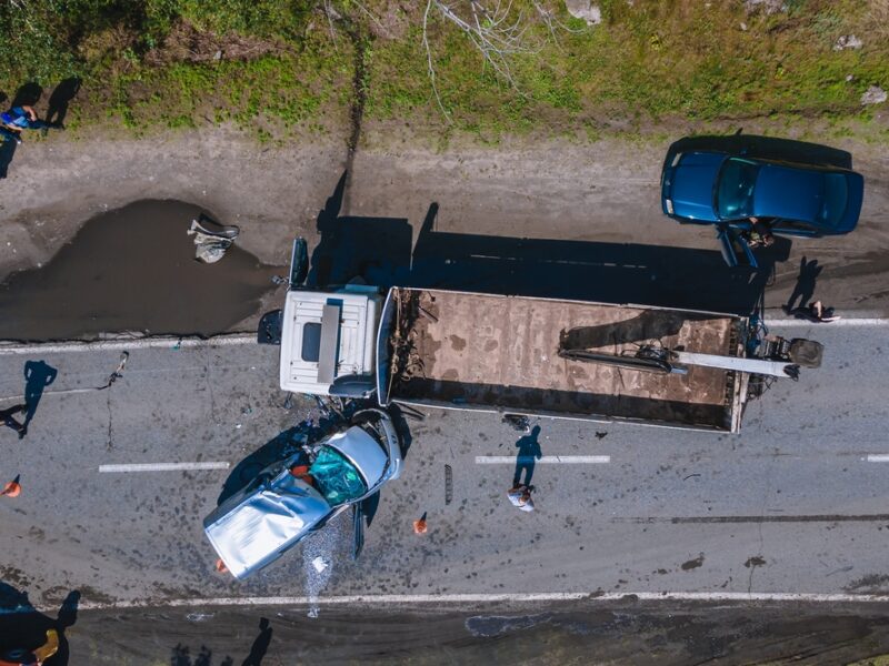 Overhead view of a 2 lane highway, a gray car has crashed into the side of a long semi truck, there are emergency personnel investigating the scene.