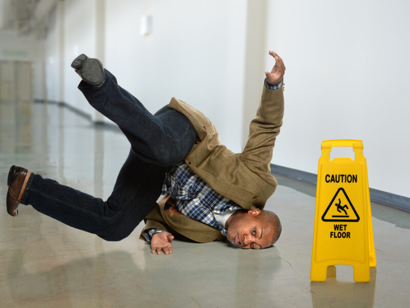 A man slips and falls next to a "wet floor" sign and hits his head on the hard surface.